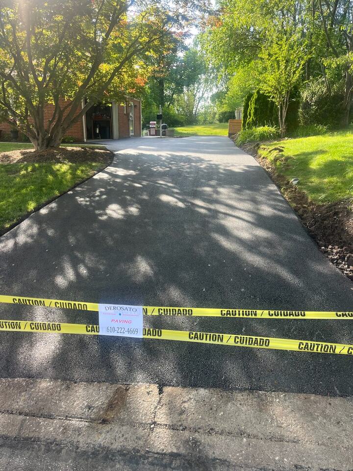 Fresh asphalt driveway in dappled shade, taped off with a DeRosato Paving sign